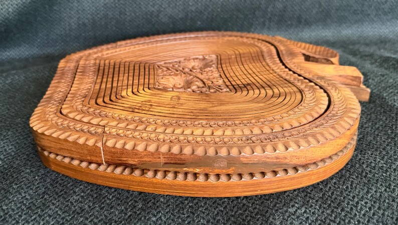May include: A wooden, collapsible fruit basket with a natural finish. The basket is carved with a floral design in the center and a decorative border. The basket is approximately 10 inches in diameter.