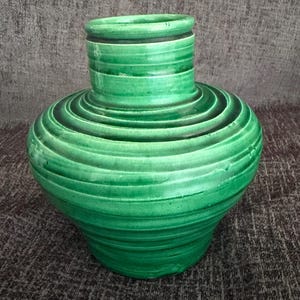 May include: A vibrant green ceramic vase featuring a wide, rounded body that tapers toward a narrow, cylindrical neck. The surface is decorated with horizontal, concentric ridges that create a textured, ribbed appearance throughout the piece.