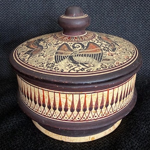 May include: A round, lidded ceramic jar with a dark brown exterior and a cream-colored interior. The lid features a detailed illustration of birds and floral patterns in black and red. The jar's body is decorated with a repeating pattern of teardrop shapes.