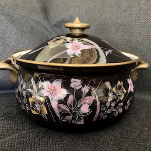 May include: A black enamel pot with a matching lid, featuring two side handles and a central lid knob in a muted gold tone. The exterior is decorated with a floral pattern of pink and white lilies, along with green and gold leaves and stems.
