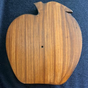 May include: A wooden apple-shaped cutting board with a stem and leaf detail. The board is made of light brown wood with vertical grain lines. A small hole is visible near the center of the board.