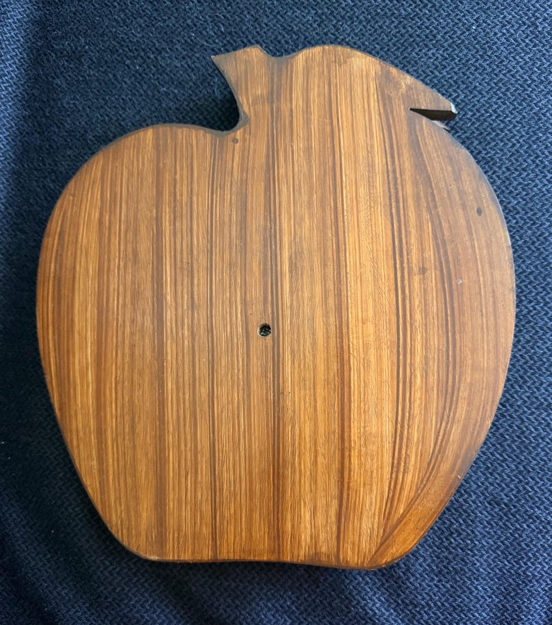 May include: A wooden apple-shaped cutting board with a stem and leaf detail. The board is made of light brown wood with vertical grain lines. A small hole is visible near the center of the board.