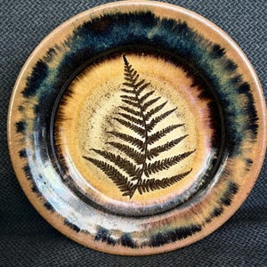 May include: A ceramic plate with a fern leaf design. The plate features a brown fern leaf in the center, surrounded by a tan and brown background. The rim of the plate has a dark blue and brown glaze. The plate is round.