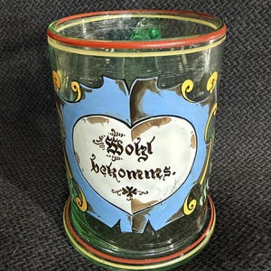 May include: A vintage glass with a heart-shaped design and the words "So ist bekommst." The glass features a blue heart with a white center, surrounded by green, yellow, and red accents. The glass is cylindrical with a flared base.