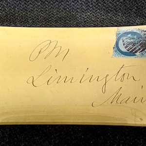 May include: A rectangular, cream-colored envelope with handwritten script and a blue postage stamp. The script reads "PM Limington Maine". The envelope is slightly curved and appears aged.
