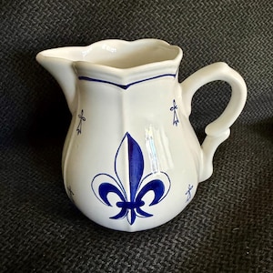 May include: A white ceramic pitcher featuring a prominent blue fleur-de-lis design on the front. The pitcher has a scalloped rim, a curved handle, and small blue ermine spot motifs scattered across the body with a blue line near the top rim.