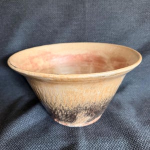 May include: A ceramic bowl with a wide, flared rim. The exterior features a gradient color scheme, transitioning from a light tan and beige upper section to a dark, textured charcoal and black base. The interior has a smooth, light tan finish.