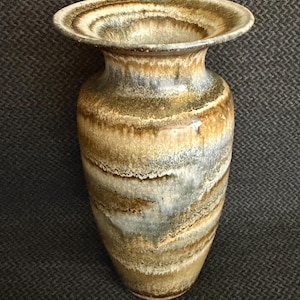 May include: A ceramic vase featuring a wide, flared rim and a tapered body. The surface displays a reactive glaze with horizontal bands of earthy brown, tan, and muted gray tones, creating a layered, mottled appearance across the entire exterior.