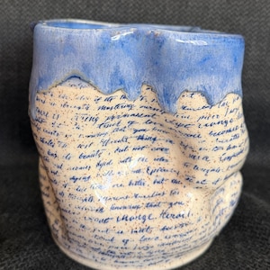 May include: A handmade ceramic mug with a unique design. The top rim is a light blue, with the rest of the mug a speckled off-white. The surface is covered in blue handwritten text. A unique piece for coffee or tea.