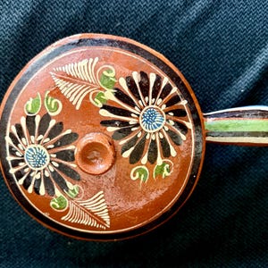 May include: A round, ceramic pot with a lid and handle. The pot is brown with black, white, green, and blue floral designs. The handle has green, white, and black stripes. The pot is likely used for cooking or serving.