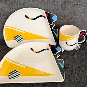 May include: Two off-white ceramic plates with a semi-circular shape and a matching mug. The pieces feature a yellow triangular graphic, a black wavy line with colorful segments, and a circular pattern with black, white, and teal stripes.