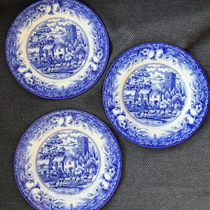 May include: Three blue and white decorative plates. Each plate features a central village scene with a church and figures, surrounded by a floral and wheat pattern border. The plates are arranged on a gray surface.