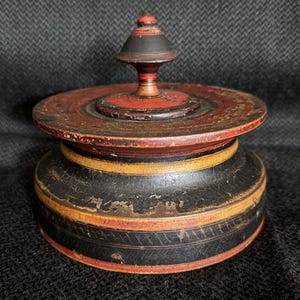 May include: A vintage, round wooden box with a lid. The box is painted with red, black, and yellow bands. The lid has a conical finial. The paint shows signs of wear, giving it a rustic appearance. The box is likely used for storage.
