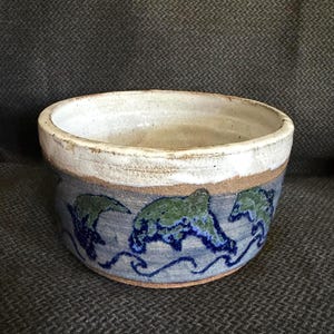 May include: A round ceramic bowl with a thick white glazed rim and a textured blue-grey base. The exterior features a repeating hand-painted pattern of green and dark blue dolphins leaping above stylized blue waves, with visible clay edges.