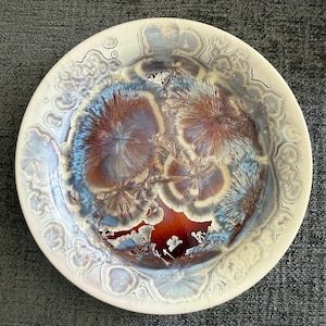 May include: A round ceramic bowl featuring a crystalline glaze with a smooth, glossy finish. The surface displays intricate, starburst-like patterns in shades of deep brown, rust, and light blue, set against a creamy off-white background.