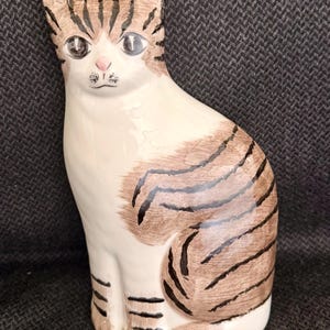 May include: A ceramic cat figurine in a seated position. The cat has a white body with brown stripes and markings. The cat's face has large eyes, a pink nose, and a small black mustache. The figurine is on a dark gray background.