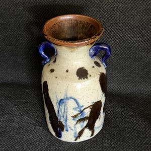 May include: A ceramic vase with a speckled cream-colored body, featuring two blue handles and a brown rim. The exterior is decorated with dark brown and blue abstract brushstroke patterns, creating a textured appearance on the cylindrical vessel.