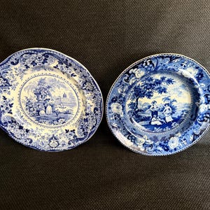 May include: Two round ceramic plates featuring blue and white transferware patterns. Each plate displays a central landscape scene with figures and buildings, surrounded by a wide border of intricate floral and scrollwork designs.
