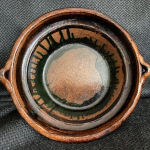 May include: A handmade, round ceramic bowl with a dark brown exterior and a green and copper-colored interior. The bowl has two small handles and a rustic, textured appearance, suggesting it is a piece of pottery.