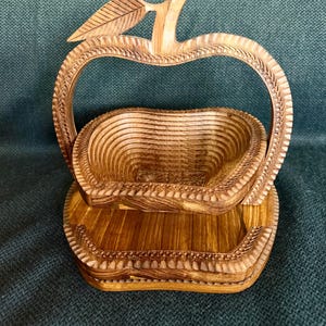 May include: A wooden apple-shaped basket with a handle and a base. The basket is made of light brown wood and has a carved design around the edges. The handle is shaped like the top of an apple with a leaf.