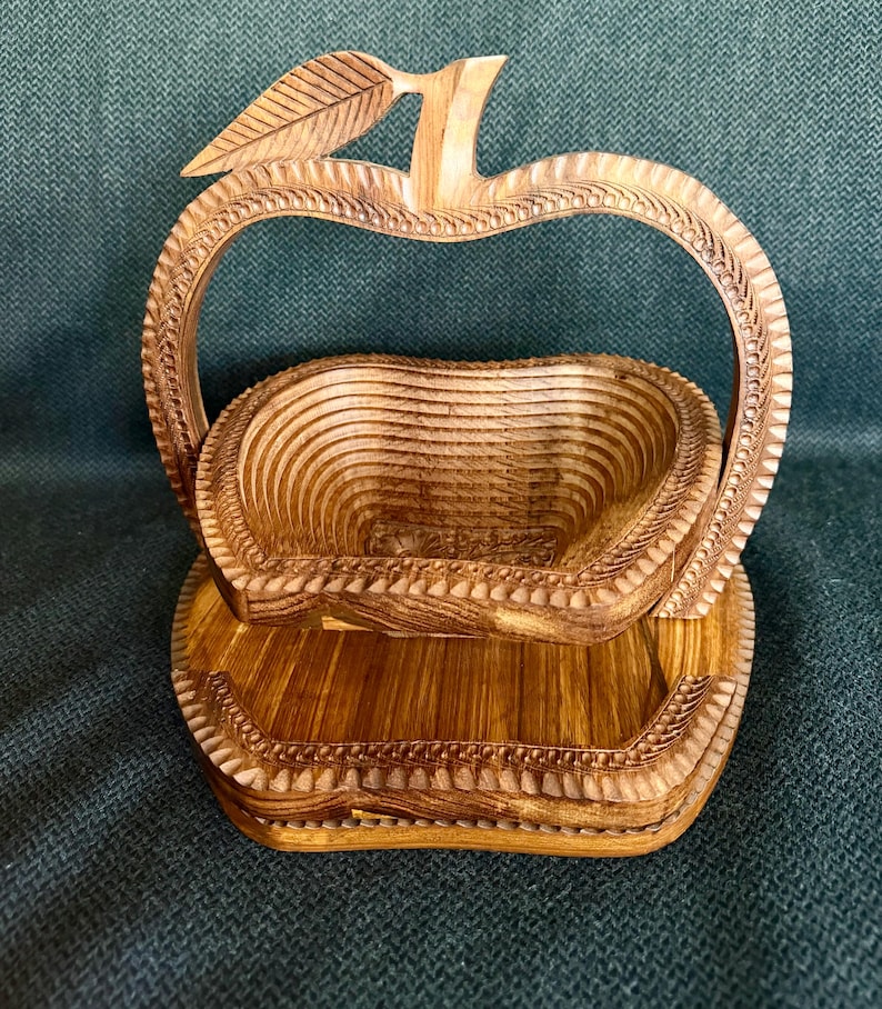 May include: A wooden apple-shaped basket with a handle and a base. The basket is made of light brown wood and has a carved design around the edges. The handle is shaped like the top of an apple with a leaf.