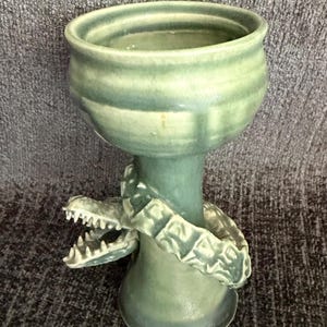 May include: A sage green ceramic goblet featuring a sculpted alligator wrapped around the stem. The alligator has a textured, scaly body and an open mouth with visible teeth. The goblet has a smooth, glossy finish and a wide, circular rim.
