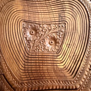 May include: A wooden fruit basket with a carved floral design in the center. The basket is made of thin, curved wooden slats, and the edges are intricately carved with a repeating pattern. The wood has a warm, natural tone.