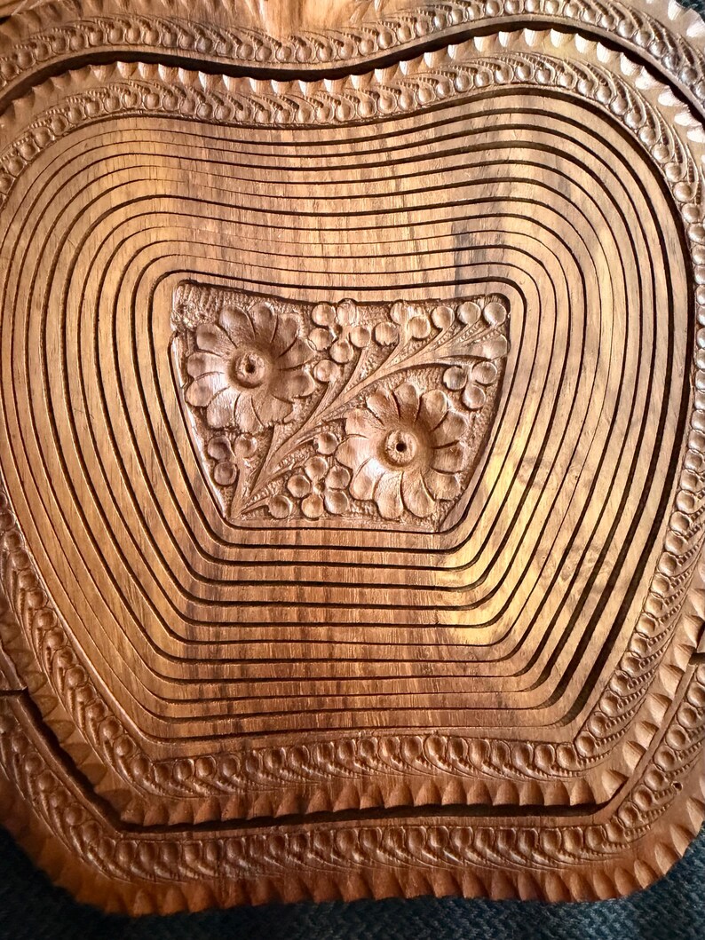 May include: A wooden fruit basket with a carved floral design in the center. The basket is made of thin, curved wooden slats, and the edges are intricately carved with a repeating pattern. The wood has a warm, natural tone.