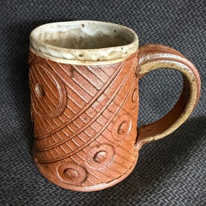 May include: A cylindrical ceramic mug with a reddish-brown exterior featuring a textured pattern of diagonal lines and raised circular motifs. The mug has a thick, curved handle and a contrasting off-white, speckled interior rim finish.