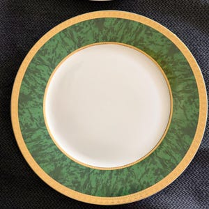 May include: White ceramic plates with a green marble-like border and gold trim. The plates have a gold inner rim and a white center. The plates are arranged on a dark blue surface.