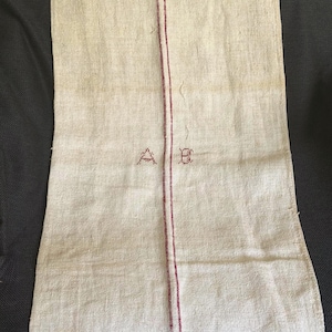 May include: A rectangular beige linen sack featuring a vertical red stripe running down the center. The letters A and B are embroidered in red thread on either side of the stripe. A white drawstring is visible at the top left corner of the sack.