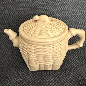 May include: A light beige ceramic teapot featuring a textured exterior that mimics the appearance of woven wicker basketry. The lid is topped with a small sculpted animal figure, and the handle and spout are shaped to resemble bamboo segments.