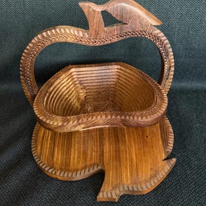 May include: A wooden fruit basket with a carved handle and base. The basket is made of light and dark brown wood and has a unique apple shape. The handle is arched and features a bird-like design at the top.