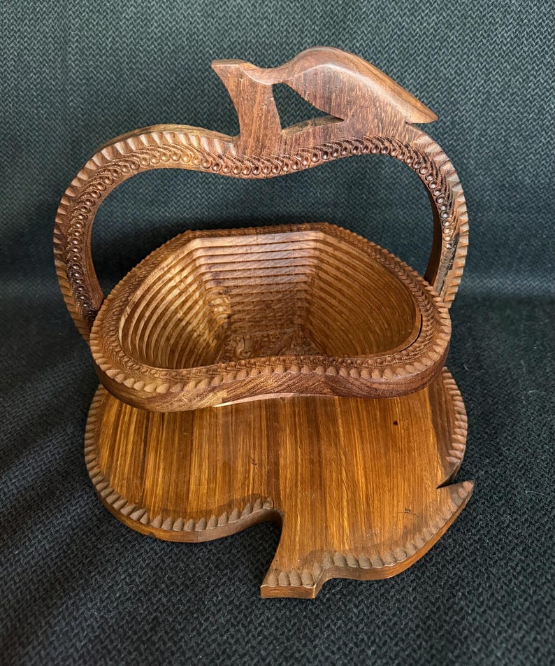 May include: A wooden fruit basket with a carved handle and base. The basket is made of light and dark brown wood and has a unique apple shape. The handle is arched and features a bird-like design at the top.