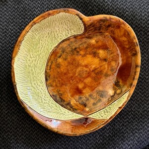 May include: A round ceramic dish featuring a dual-tone glaze. One section has a textured, pale yellow surface with fine, radiating lines, while the other section displays a smooth, glossy, mottled brown finish with dark amber and tan highlights.