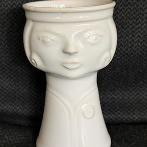 May include: A white ceramic vase with a face design. The vase has a wide top, a face with eyes, a nose, and a mouth, and a long neck. The vase is approximately 7 inches tall.