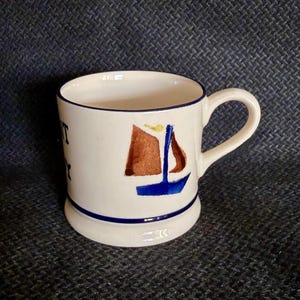 May include: A cream-colored ceramic mug featuring a hand-painted sailboat illustration with brown sails and a blue hull. The mug has a dark blue rim and a matching blue horizontal stripe near the base, with partial black text visible on the side.