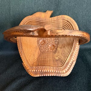 May include: A wooden apple-shaped basket with a carved handle. The basket is made of light brown wood and features intricate carved details, including floral patterns and a textured rim. The handle is curved and attached to the top of the apple.