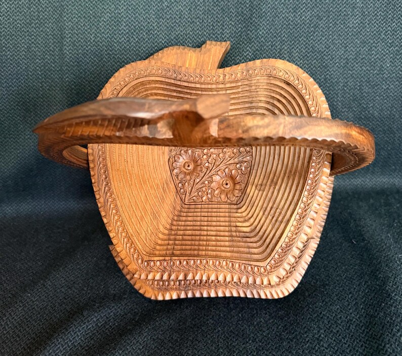 May include: A wooden apple-shaped basket with a carved handle. The basket is made of light brown wood and features intricate carved details, including floral patterns and a textured rim. The handle is curved and attached to the top of the apple.