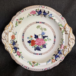 May include: A white ceramic serving platter with a scalloped edge and gold accents. The platter features a central floral design with pink, red, and blue flowers. Additional floral and foliate patterns are scattered around the rim.