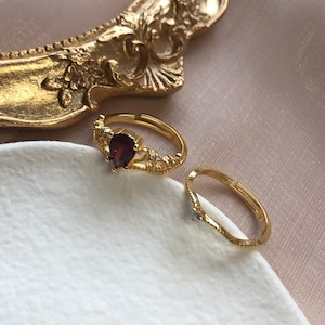 May include: Two gold rings with intricate designs. One ring features a red gemstone, while the other has a smaller clear stone.