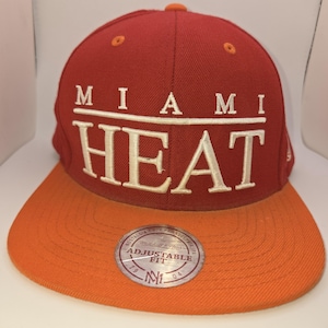 May include: A red and orange Miami Heat adjustable baseball cap. The cap features the team name in white block letters across the front. The brim is orange, and a circular patch with text is visible.