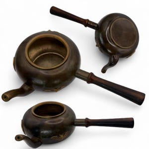 May include: Three antique bronze teapots with dark wooden handles. Each teapot has a round body, a spout, and a decorative element near the base. The teapots are arranged on a white background.