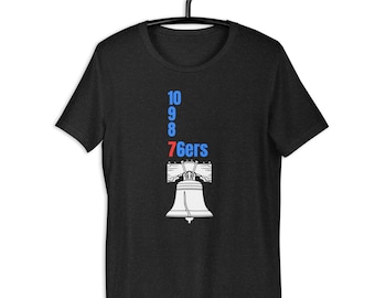 May include: Dark gray t-shirt featuring the text "10 9 8 76ers" in blue and red, with a white illustration of the Liberty Bell. The shirt is hanging on a black hanger.