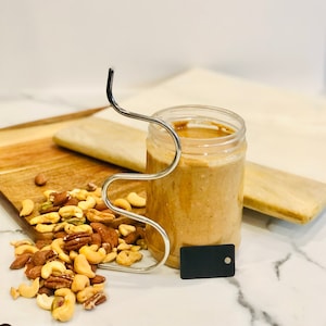 May include: A clear jar filled with light brown nut butter, held by a silver metal handle. A pile of mixed nuts sits on a wooden cutting board next to the jar. A small black tag is attached to the handle.