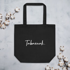 May include: Black tote bag featuring the word "Tabarnak." in white script. The bag has a rectangular shape with a handle. The background is a light grey with cotton branches. The bag is made of a sturdy material.