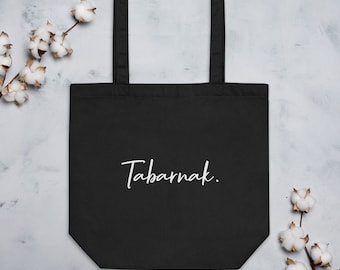 May include: Black tote bag with the word "Tabarnak." in white script. The bag has a rectangular shape with a handle and is made of a sturdy material. The background is a light gray with cotton branches.