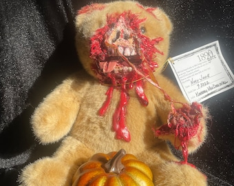 May include: A vintage teddy bear with a gruesome, bloody, and realistic-looking injury to its face and arm. The bear is sitting on a black surface with a small, golden pumpkin in front of it. A tag attached to the bear's arm reads "1890 Gore" and "Nasty Jackie 7-1-2012  Kimberly Van Der Wall".
