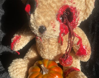 May include: A beige teddy bear with a bloody eye and a pumpkin. The bear's eye is missing and there is a red, bloody hole where the eye should be. The bear is sitting on a black surface.