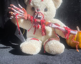 May include: A white teddy bear with red blood splatters and a bloody skeletal arm sticking out of its mouth. The bear is sitting on a black surface.